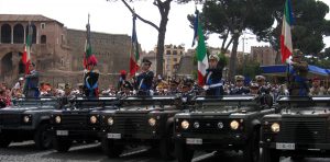 Parata militare con soldati italiani in uniforme su veicoli, sventolando bandiere tricolori, simbolo di dovere e giustizia